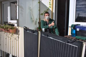 Ein junger Mann sitzt auf seinem Balkon, reckt den Daumen nach oben, im Vordergrund das neue Balkonkraftwerk.