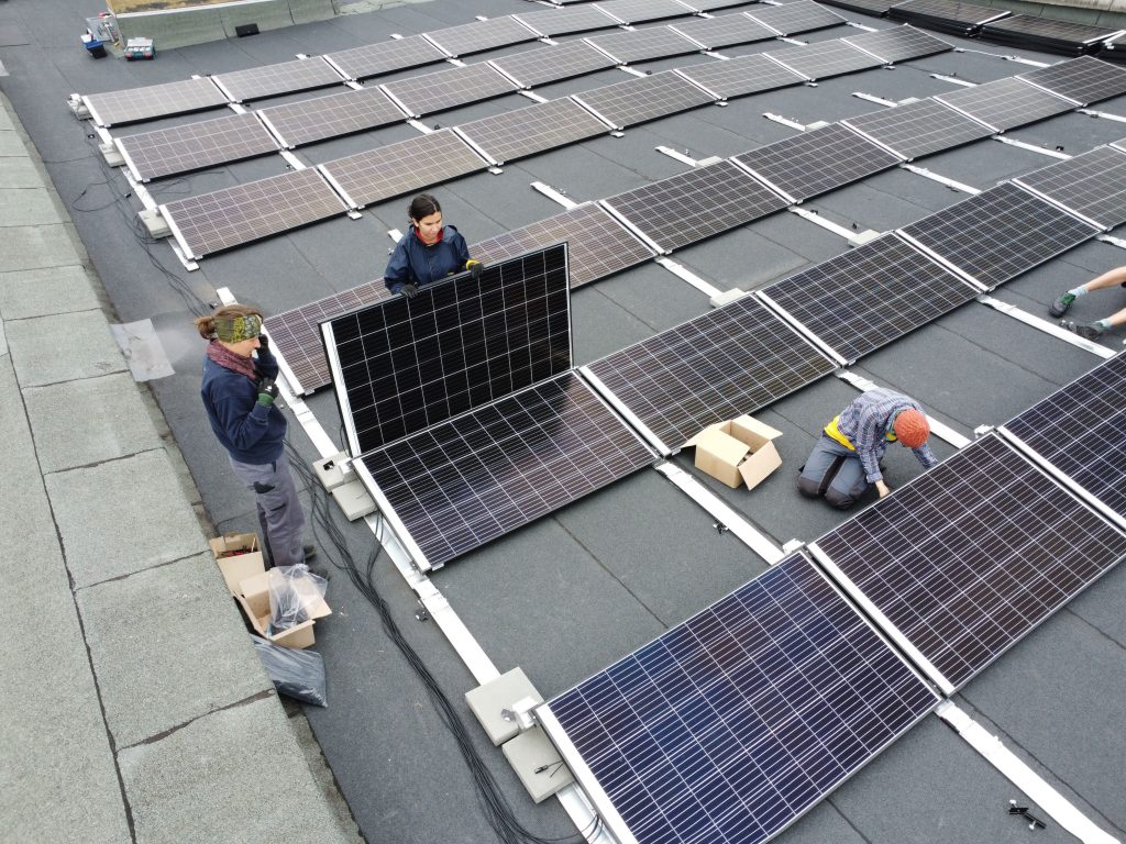 Drei Frauen auf dem Sandershaus-Dach, wo sie gerade eine Flachdach-Photovoltaik-Anlage bauen.