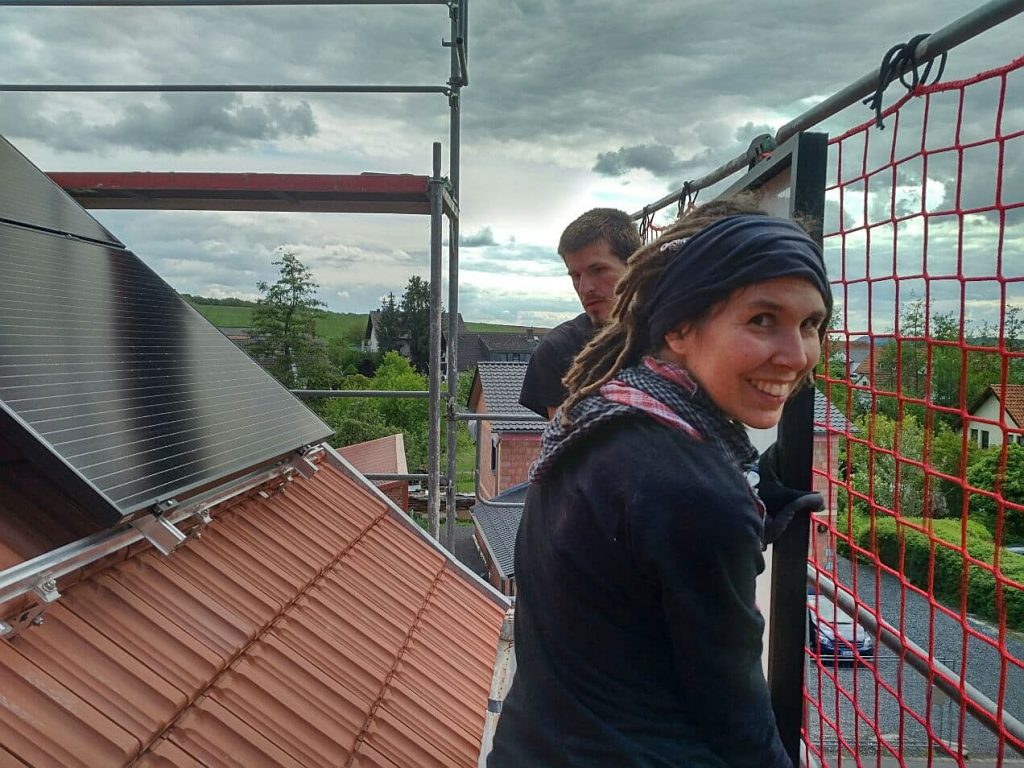 Zwei junge Personen auf einem Gerüst neben einem Dach, wo gerade eine Photovoltaik-Anlage gebaut wird.