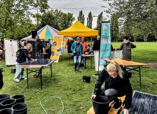 Menschen, die gebrauchte Photovoltaik-Module putzen und deren Stecker tauschen in einem Park mit gelbem Pavillon.